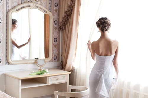 A bride facing a window, wearing a gown with a bustle inserted in the back.
