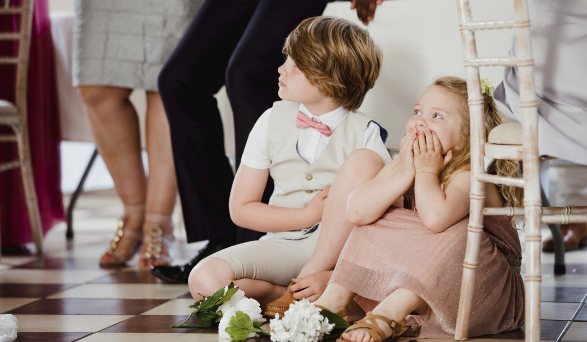 Children are sitting on the dancefloor by a table at a wedding. They are watching the bride and groom share their first dance.